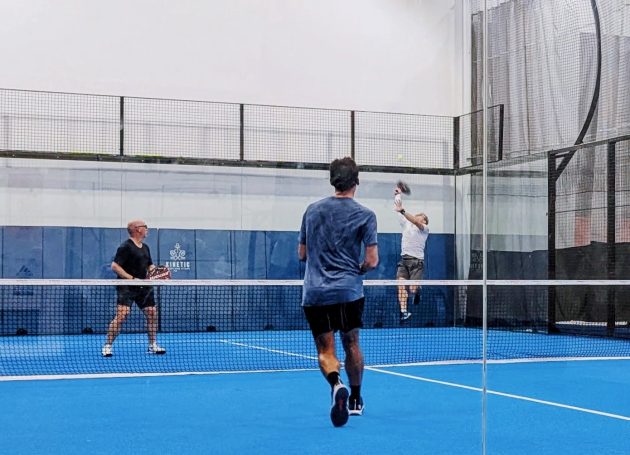 A padel player retrieves a lob over his head as his opponent rushes to the net to prepare for the next shot. 