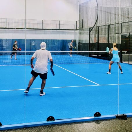 A padel player on the far side of a padel court hits a backhand lob as his partner looks on and the opponents prepare to respond. 