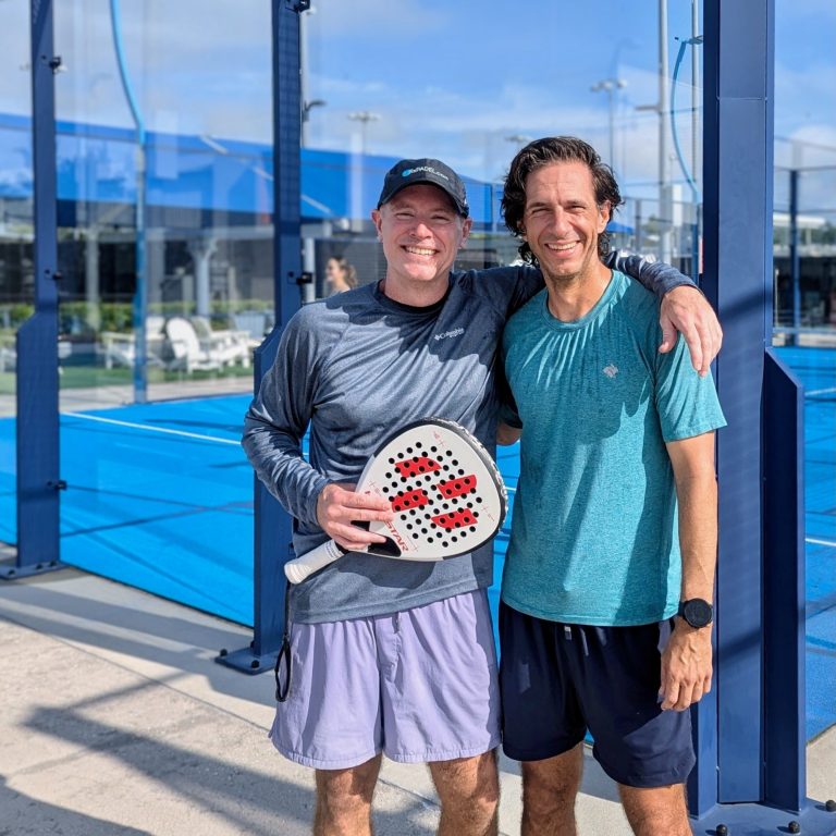 Two padel players, one holding pala or padel racquet, pause outside the court for a photo together. 
