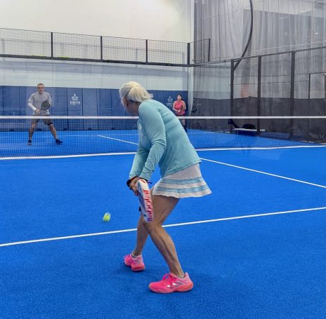 A padel player prepares a backhand return of serve as two opponents across the net watch on. 