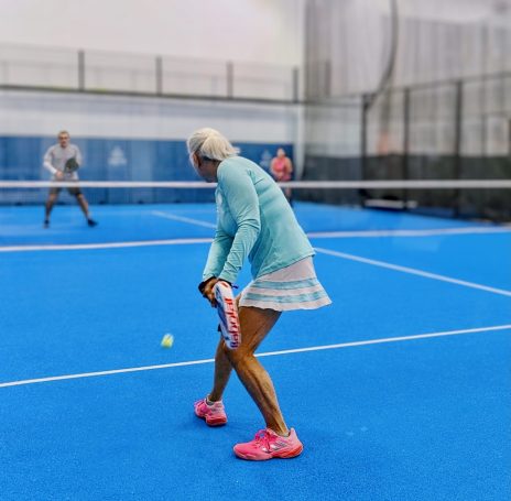 A padel player prepares a backhand return of serve as two opponents across the net watch on. 