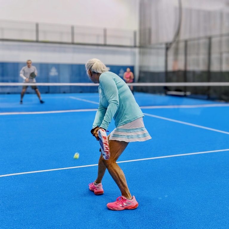 A padel player prepares to hit a backhand return of serve as two opponents across the net ready themselves for the shot. 