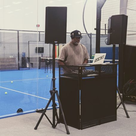 A music dj plays a set with a blue padel court in use behind him
