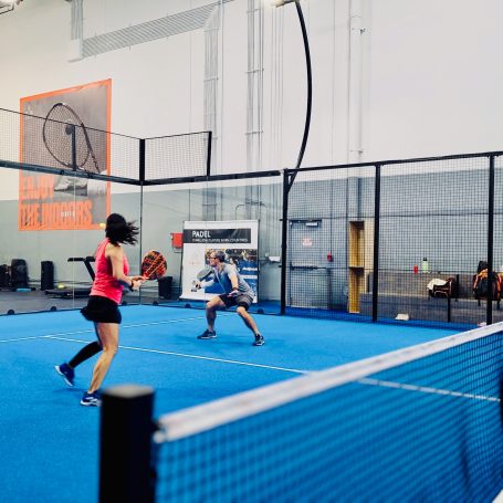 A man and a woman work to return a shot off the back wall of a blue padel court.