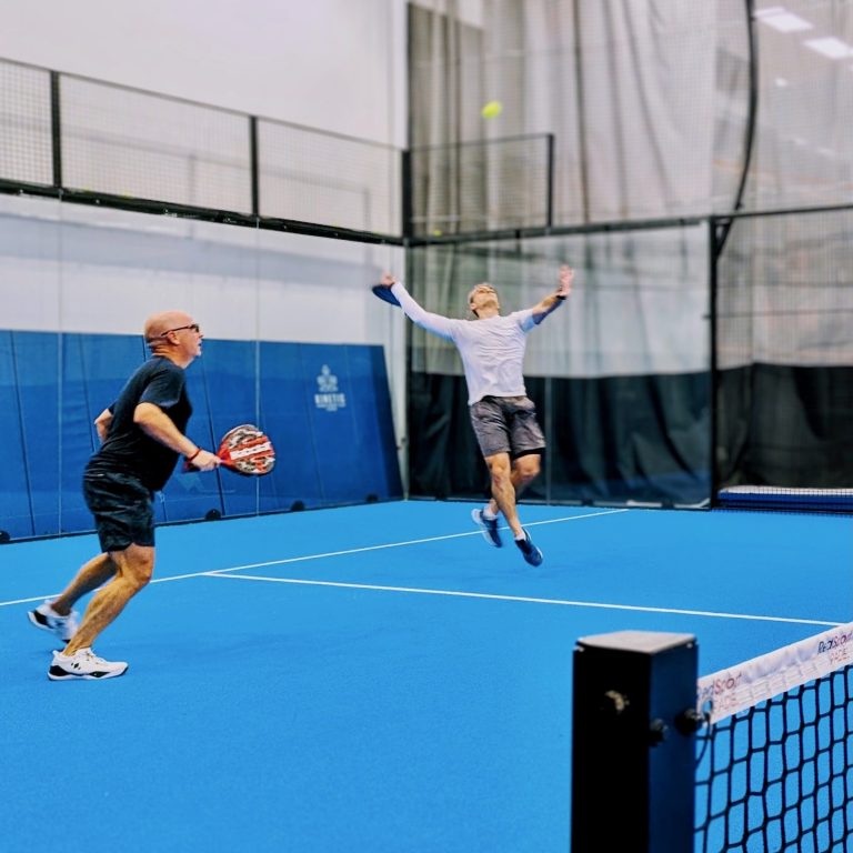 A padel player leaps off the ground to hit a ball that was lobbed over his head while his partner prepares for the next shot. 